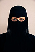 Close-up portrait of woman wearing black muslim hijab and muslim dress, looking at camera, studio shot on white background