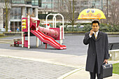 Geschäftsmann mit Kinderregenschirm auf dem Spielplatz