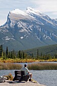 Alberta, Kanada; Ein männlicher Wanderer sitzt auf einer Bank mit Blick auf den Mount Rundle und die Vermilion-Seen
