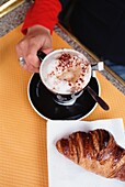 Frau im Café mit einer Tasse Cappuccino und Croissant im Vordergrund