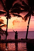 Einheimische in einem Grasrock hält eine brennende Fackel an der Küste bei Sonnenuntergang