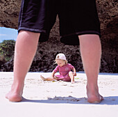 Blick durch die Beine des Vaters auf ein Kind, das im Sand spielt