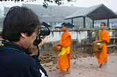 Photographer Taking Pictures Of Novice Monks Out Collecting Alms At Dawn.