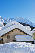 Schneebedeckte Dächer von Häusern mit schroffen, schneebedeckten Bergen im Hintergrund; San Bernardino, Graubünden, Schweiz