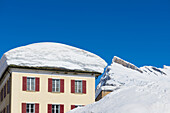 Schneehaufen auf einem Dach mit schneebedeckten Bergen im Hintergrund und blauem Himmel; San Bernardino, Graubünden, Schweiz