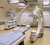 A modern hospital room, a large portable mobile scanning machine with curved shaped arms, a mobile scanner and hospital gurney or bed. 