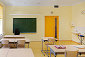 Klassenzimmer mit Schreibtisch und Stühlen. Fenster mit gelben Jalousien und grüner Tafel.