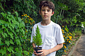 Ein Junge hält einen kleinen Baumschössling in einem Topf, der in einem Garten steht. 