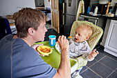 Father feeding toddler in high chair in kitchen