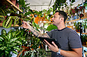 Mann mit Tattoos, der in einem Blumenladen arbeitet und den Bestand mit einem Tablet überprüft
