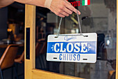 A person hanging up a sign on a restaurant door, reading Close. Closed, dual language, English and Italian.