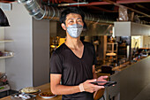 Ein Mann, der in einem Restaurant arbeitet, trägt eine Gesichtsmaske, steht vor einer offenen Küche und hält ein digitales Tablet in der Hand. 
