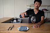 A man pouring red wine into a wine glass, a place setting on a restaurant counter. 