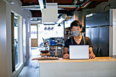 Ein Mann mit einer Gesichtsmaske in einer Restaurantküche, der einen Laptop benutzt, der Besitzer oder Manager. 