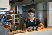 Ein Mann mit einer Gesichtsmaske an der Theke einer Restaurantküche, der ein digitales Tablet benutzt, der Eigentümer oder Manager. 