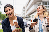 Mittlere Einstellung von zwei Unternehmerinnen, die lachen und sich über Inhalte in den sozialen Medien freuen, während sie im Freien arbeiten