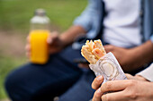 Medium shot of a hand holding a wrapped sandwich and an out-of-focus hand holding an orange juice bottle