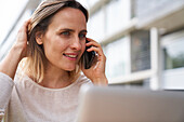 Porträt einer Frau, die vor einem Laptop sitzt und mit einem Mobiltelefon spricht