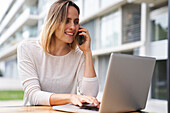 Porträt einer Frau, die im Freien an einem Laptop-Computer arbeitet und dabei mit einem Mobiltelefon spricht