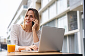 Porträt einer amüsierten Frau, die im Freien mit einem Laptop-Computer sitzt, während sie mit einem Mobiltelefon spricht und Orangensaft trinkt