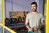 Mittlere Einstellung eines lächelnden, bärtigen Mechanikers, der in seiner Werkstatt vor seinen Werkzeugen steht