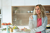 Porträt einer attraktiven Frau mittleren Alters, die eine Tasse Kaffee hält und in die Kamera schaut, stehend in der Küche