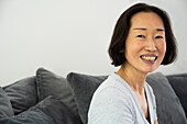 Porträt einer asiatisch-amerikanischen Seniorin, die lächelt und in die Kamera schaut, während sie im Wohnzimmer sitzt