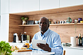 Älterer afroamerikanischer Mann sitzt in der Küche und isst Obst zum Frühstück, während er Nachrichten auf einem digitalen Tablet abruft
