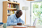 Älterer afroamerikanischer Mann mit Kopfhörern, während er zu Hause an einem Laptop-Computer arbeitet