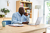 Afroamerikanischer Mann mittleren Alters, der ein Telefonat führt, während er zu Hause mit einem Laptop arbeitet