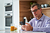 Berufstätiger Mann mittleren Alters, der beim Frühstück auf der Küchentheke zu Hause Nachrichten auf dem Telefon abruft