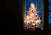 Beleuchteter weißer Weihnachtsbaum vor einem nächtlichen Hausfenster