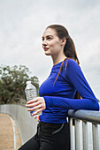 Lächelnde Frau in Sportkleidung, die sich an einen Zaun lehnt und eine Flasche Wasser hält
