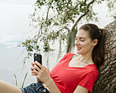 Lächelnde Frau lehnt an einem Ast und schaut auf ihr Smartphone