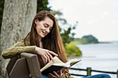 Lächelnde Frau liest ein Buch auf einer Bank am Fluss
