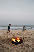 Boy (8-9) and girl (2-3) playing on beach near bonfire