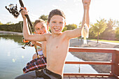 Portrait of smiling shirtless boy (8-9) holding fish and fishing rod by lake