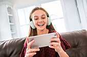 Lächelndes Mädchen (12-13) mit Kopfhörern und Smartphone auf dem Sofa