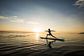 Frau bei der Ausführung von Virabhadrasana II (Krieger-II-Pose) auf dem Paddleboard bei Sonnenuntergang