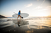 Frau in blauem Badeanzug trägt Paddleboard auf See bei Sonnenuntergang