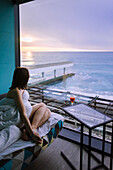 Frau auf dem Bett sitzend mit Cocktail im Hotel und Sonnenaufgang über dem Meer betrachtend