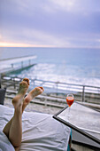 Frauenfüße und Cocktail im Hotelzimmer mit Blick auf das Meer bei Sonnenaufgang