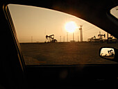 Silhouette von Pumpenhebern auf einem Ölfeld bei Sonnenuntergang vom Auto aus gesehen