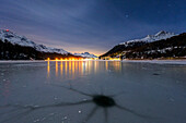 Löcher im gebrochenen Eis auf dem Champfer See mit dem Dorf Silvaplana im Hintergrund bei Nacht, Engadin, Graubünden, Schweiz, Europa