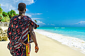 Maasai-Mann mit traditioneller Kleidung und Stock bewundert das kristallklare Meer an einem Strand, Sansibar, Tansania, Ostafrika, Afrika