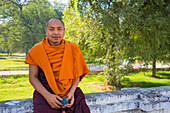 Mönch in safranfarbener Robe, der mit seinem Mobiltelefon in der Hand an der Wand sitzt, Königspalast, Mandalay, Myanmar (Burma), Asien