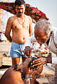 Baeber shaving the head and beard of a pilgrim, Dashashwamedh ghat (main ghat), in Ganges river, Varanasi, Uttar Pradesh, India.