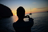 Silhouette einer jungen Frau, die den Sonnenuntergang und ein Segelboot am Strand fotografiert