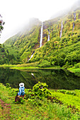 Rear view of woman with blue cloth and white hat that sits and admires the Ribeira do Ferreiro waterfall among green and lush vegetation with clouds, Poco Ribeira do Ferreiro (Alagoinha), Lajes das Flores municipality, Flores Island (Ilha das Flores), Azores archipelago, Portugal, Europe