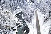 Car traveling on mountain road crossing the forest covered with snow and frozen river, aerial view, Switzerland